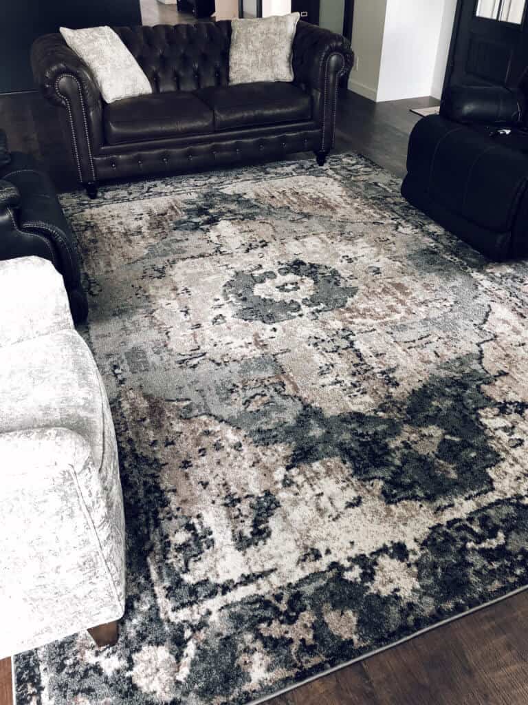 Large area rug with worn industrial vintage patterns in colors of Charcoal, Dark Brown, Camel, Medium Gray, White surrounded by chesterfield furniture and barrel chairs