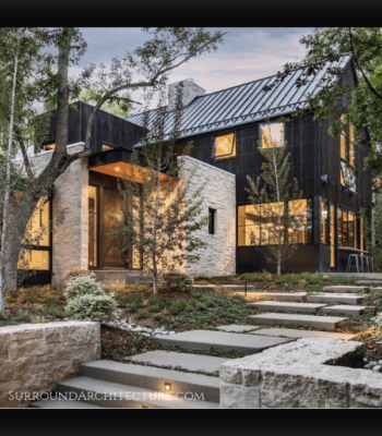 Dream House exterior features modern lines in the dark vertical siding mixed with rustic light white stone nestled in the trees with stone landscape surrounding it